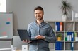 © Prostock-studio - Smiling Caucasian Man In His 30s Holding Laptop While Standing In Modern Office Setting, Engaging With Technology And Working On Professional Tasks, Showcasing Business Concept.