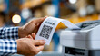 © Nataliya - Faceless hand holding printed receipt with QR code emerging from thermal printer in warehouse, barcode printing concept, logistics photography, blurred warehouse background, with