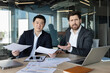 © Liubomir - Two diverse business colleagues sitting at an office desk, holding papers and expressing confusion and stress while analyzing financial documents and discussing a challenging situation