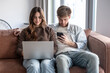© luciano - Young couple using laptop and smartphone at home, sitting on sofa, connecting with digital devices, browsing internet, working remotely, and enjoying free time together