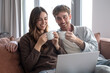 © luciano - Happy young couple enjoying morning coffee and laughter together on a cozy sofa at home, sharing relaxing moments while watching online content on a laptop