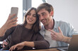 © luciano - Happy young adult couple smiling while taking a selfie together with a smartphone, sharing a joyful moment and connecting remotely from their home