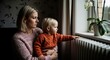 © Hungarian - Concerned Mother and Toddler Sitting by Radiator Looking Out Window During Winter Energy Crisis