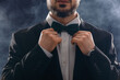 © Pixel-Shot - Young man in stylish suit adjusting bow tie on smoke background, closeup