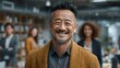 © Anuson - A happy and confident middle-aged Asian businessman with a beard smiling brightly in a modern office with blurred colleagues in the background