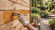 © Pavel - Hand in glove applying protective varnish or stain with brush to wooden fence boards outdoors in sunny backyard.