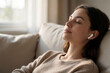 © Maryna - Young woman listens to music with wireless earbuds, resting on a comfortable sofa at home. She smiles with eyes closed, enjoying a moment of peace and quiet relaxation on the couch.