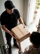 © Maryna - Delivery courier in black uniform hands cardboard package to customer at doorway. Worker brings order to home for recipient. Person receives parcel from shipping service employee at front door. Fast