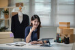 © BTM_Graphic - Young Asian Woman Entrepreneur Working at Home Office with Laptop and Shipping Boxes for Online Fashion Business