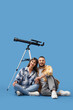 © Pixel-Shot - Young couple with telescope sitting on blue background