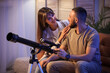 © Pixel-Shot - Young couple with telescope sitting on sofa in room at night