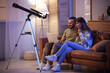 © Pixel-Shot - Young couple using laptop on sofa and telescope in room at night