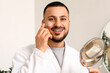 © Pixel-Shot - Young man with mirror applying cream on face in bathroom, closeup