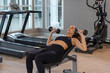 © Ron - oung modern woman training with dumbbells on an exercise bench in a bright gym, combining strength workout, stretching and mindful focus for a healthy balanced lifestyle.