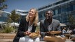 © Alexei - Caucasian woman and African American man enjoying lunch outdoors at a modern office complex, holding takeout boxes with greenery and glass buildings in the background