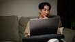 © wattana - Smiling man using a laptop while relaxing on a comfortable sofa in a warm living room at night.