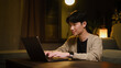 © wattana - Young man working on a laptop at home during the evening in a warm and comfortable living room.