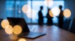 © CuteBee - Closed laptop computer resting on wooden desk with silhouettes of business professionals having a meeting in blurred office interior during soft golden hour light