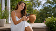 © Vyatcheslav - Smiling young woman using smartphone on wooden porch steps. Happy female in white summer dress texting outdoors. Mobile communication concept