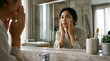 © T57 Media - Young Asian woman looking at her reflection in the bathroom mirror while performing a daily skincare routine and touching her face