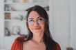 © Daniel - Young brunette woman wearing stylish glasses and an orange top, smiling confidently at the camera while standing in a bright home interior with a bookshelf in the background