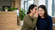 © Xenia - Young Chinese woman whispering a secret to her friend. Two smiling Asian female friends gossiping and sharing a funny story in a modern indoor setting