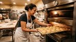 © Adnan - Smiling female baker in white apron sliding metal baking sheet with golden sesame seed burger buns into commercial oven in busy bakery kitchen