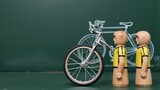 Two wooden cyclists stand beside a bike, with a chalk drawing in the background.