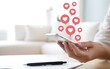 © MDRAFIQUL - Person holding smartphone with red heart bubble icons floating above screen in bright room.