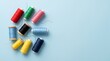 © GunGun - Top-down flat lay of colorful sewing thread spools arranged on the left side of a smooth pastel blue background, featuring vibrant red, green, pink, black, sky blue, yellow, and navy blue threads.