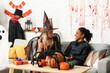 © Pixel-Shot - Cute happy girls in different costumes dressed for Halloween party with gift box and pumpkins sitting on sofa at home