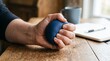 © Felippe Lopes - Close up of a person's hand squeezing a blue stress ball on a wooden table with a notebook and pen in the background