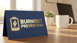 © ViskBx - Burnout prevention concept with a low battery icon on a blue card placed on a wooden office desk with a coffee mug and laptop in the background