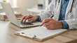© Amena - Doctor using laptop and writing on clipboard. Physician working with medical records at desk.