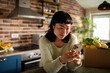 © Davor - Woman using smartphone beside grocery box in home kitchen