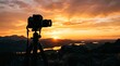 © Dara - A black camera is mounted on a tripod, capturing a vibrant sunset over a mountainous landscape. The photographer is positioned on a rocky outcrop, silhouetted against the colorful sky.