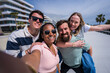 © CarlosBarquero - Four friends smile together for a selfie with an outstretched arm, modern buildings and palm trees behind them.