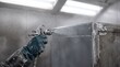 © maxici - Industrial worker applies gray primer/paint to a metal box with a spray gun. Shows the metal finishing process in a manufacturing paint booth for protective coating.