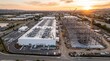 © Iryna - High Aerial of a Finished White Data Center Hall Beside a Steel Frame Building Under Construction With Four Tower Cranes Against a Hazy Golden Sunset Over Flat Farmland Industry