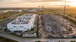© Iryna - High Aerial of a Finished White Data Center Hall Beside a Steel Frame Building Under Construction With Four Tower Cranes Against a Hazy Golden Sunset Over Flat Farmland Industry