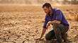 © Alena - climate impact on agriculture. Male farmer kneeling on cracked dry soil in a drought-affected field, examining the land with a concerned expression under a hazy sky