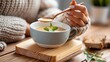 © CreativeArt - Hand in cozy sweater holding spoon, eating warm creamy soup from a bowl on wooden background, comfort food concept