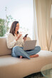 © gzorgz - Attractive woman sitting comfortable on the couch and smiling while using mobile phone and laptop, home office