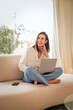 © gzorgz - Attractive woman sitting comfortable on the couch and smiling while using mobile phone and laptop, home office