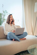 © gzorgz - Attractive woman sitting comfortable on the couch and smiling while using mobile phone and laptop, home office
