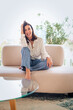 © gzorgz - Full length of a cheerful smiling confident woman sitting on the couch in front of a glass wall
