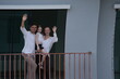© ultramansk - Caucasian family of three embracing wave happily from their balcony, the girl riding piggyback on the balcony of their new house overlooking the residential estate, authentic everyday scene