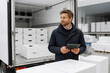 © Anastasiia - A man in a dark coat is looking at a tablet inside the open back of a white delivery truck filled with stacked boxes.