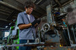 © ultramansk - Middle aged Asian small business owner in the family owned factory, dad stands at vintage green gear-hobbing machine, both black-gloved hands, skilled work of a family enterprise founder