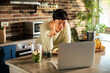 © Davor - Woman making smoothie during video call in home kitchen